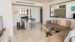 A modern living room with a glass coffee table, a sofa with patterned cushions, a TV, and a kitchen area with a dining table and chairs. - Design Apt Parking Included 2BDR TL30 (Tel Aviv)