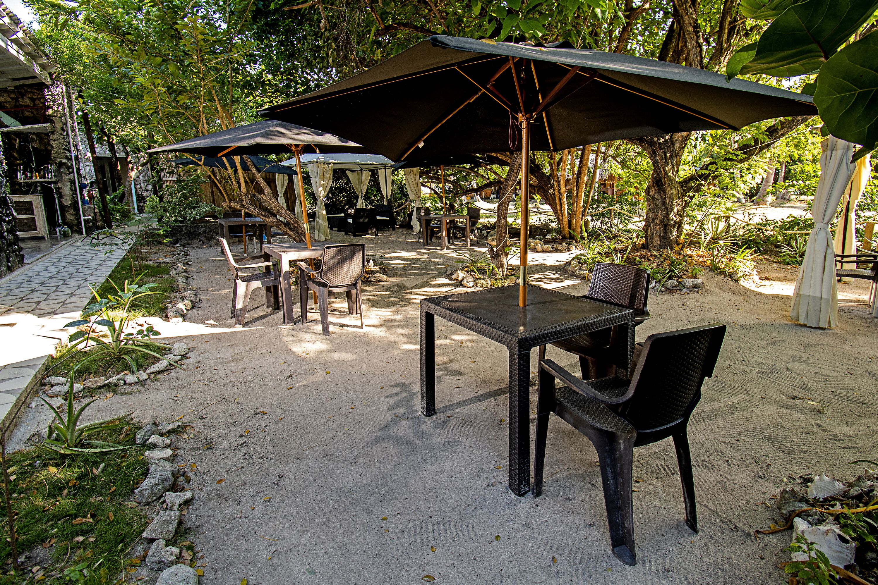 An outdoor patio with tables and chairs under large umbrellas, surrounded by trees and plants.
