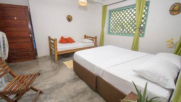 Quarto com cama de madeira, uma cama menor, uma cadeira de madeira, um ventilador e uma janela com cortina verde.