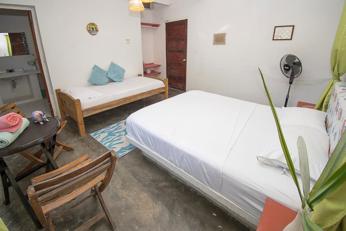 A small, clean room with two single beds, a desk, a chair, a small table, and a fan.
