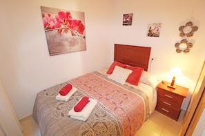 Un lit bien fait, recouvert d’une couvre-lit à motifs, avec deux serviettes rouges, une table de chevet en bois surmontée d’une lampe, et des œuvres d’art murales comprenant une peinture florale et deux autres tableaux plus petits.