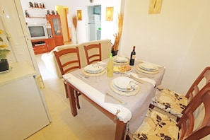 Un coin repas avec une table dressée pour quatre personnes, des chaises, un téléviseur en arrière-plan et une cuisine visible à travers une porte ouverte.
