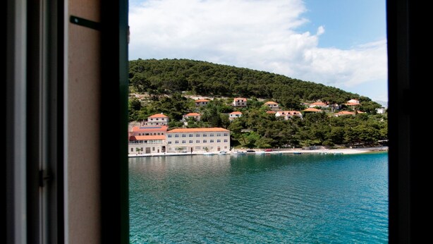 A coastal view through a window, featuring a clear blue sea, a sandy beach, and a hillside with buildings.