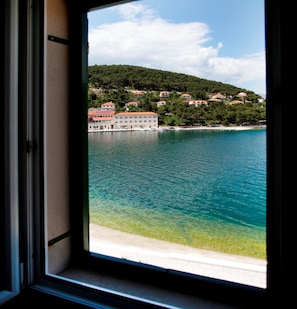 Blick durch ein Fenster auf die Küste, mit blauem Meer, Sandstrand und einer Hügelside mit Gebäuden.