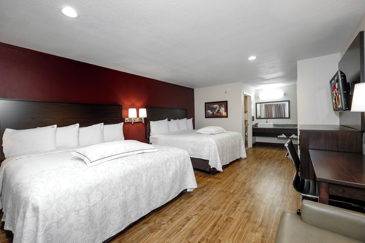 A hotel room with two beds, a desk, a chair, a TV, and a bathroom visible through an open door.