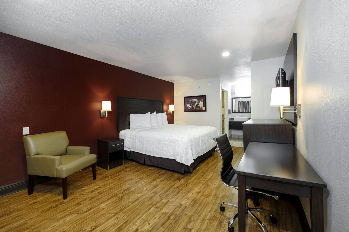A hotel room with a bed, a desk, a chair, a nightstand, and a lamp.