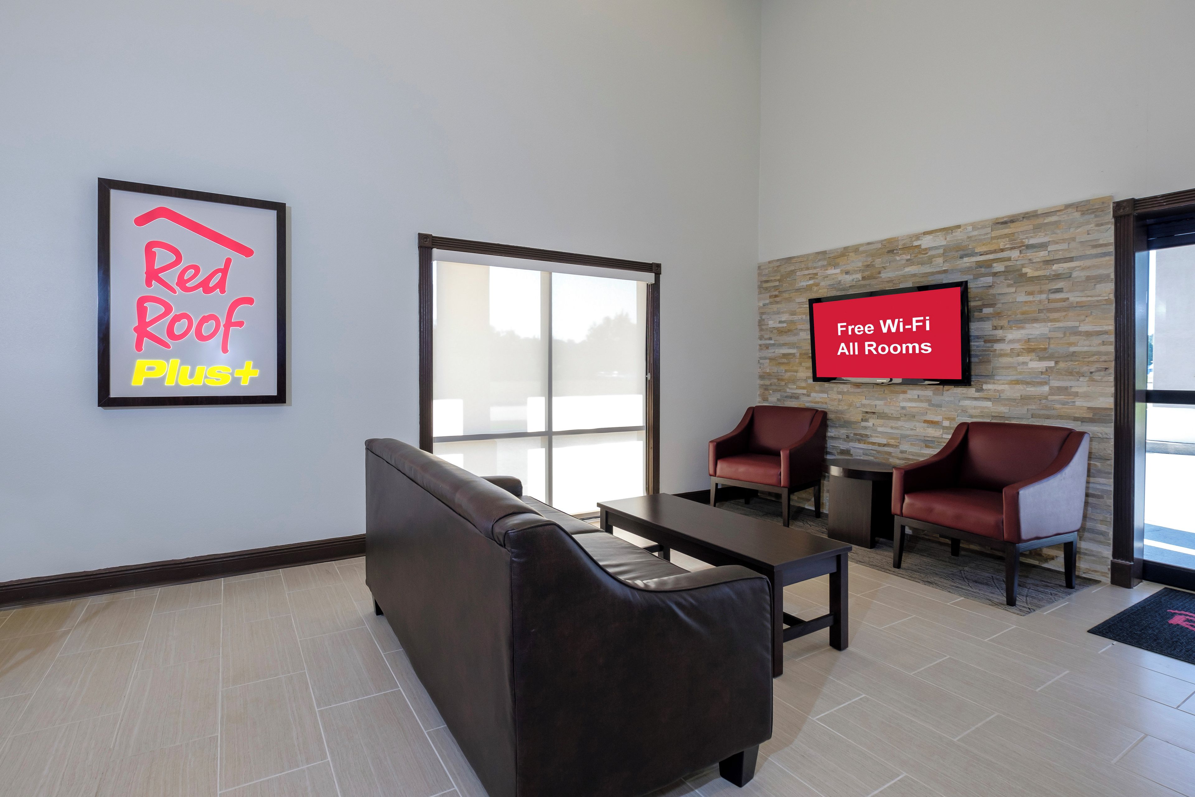 Een moderne lobby met een rood dak plus-teken, een televisiescherm waarop gratis Wi-Fi beschikbaarheid wordt weergegeven, en een zithoek met lederen stoelen.