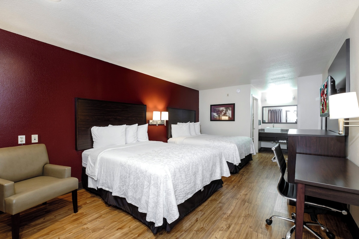 A hotel room with two beds, a desk, a chair, and a bathroom visible through an open door.