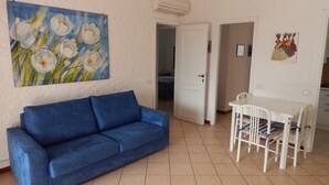 Quarto com sofá azul, mesa de jantar com cadeiras, pintura de flores e porta que dá para outro cômodo.