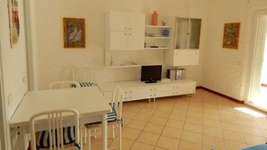Um quarto pequeno e bem iluminado, com uma mesa branca, cadeiras, uma televisão e um armário com prateleiras.