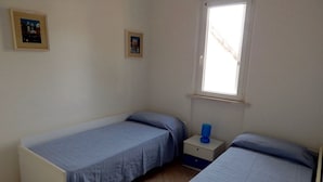 Quarto pequeno e individual com duas camas, janela e mesa de cabeceira com abajur.