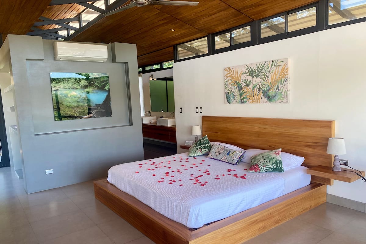 Un dormitorio moderno con estructura de cama de madera, un ventanal grande y un cuadro de plantas tropicales.