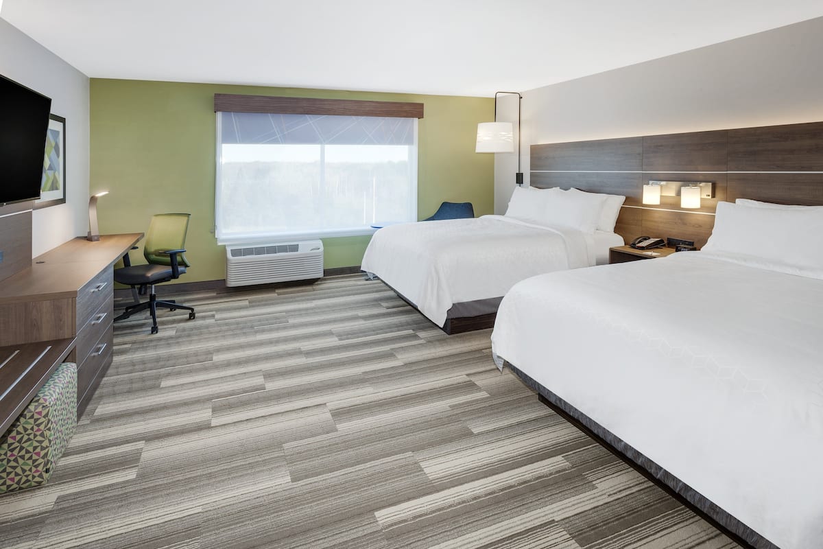 A hotel room with two beds, a desk, a chair, a TV, and a window with an air conditioner.