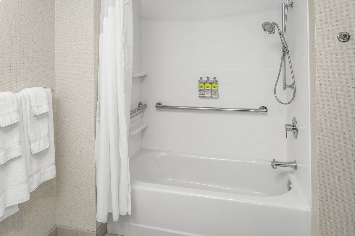 A white bathtub with a showerhead and a grab bar. There are three bottles of shampoo on the wall.