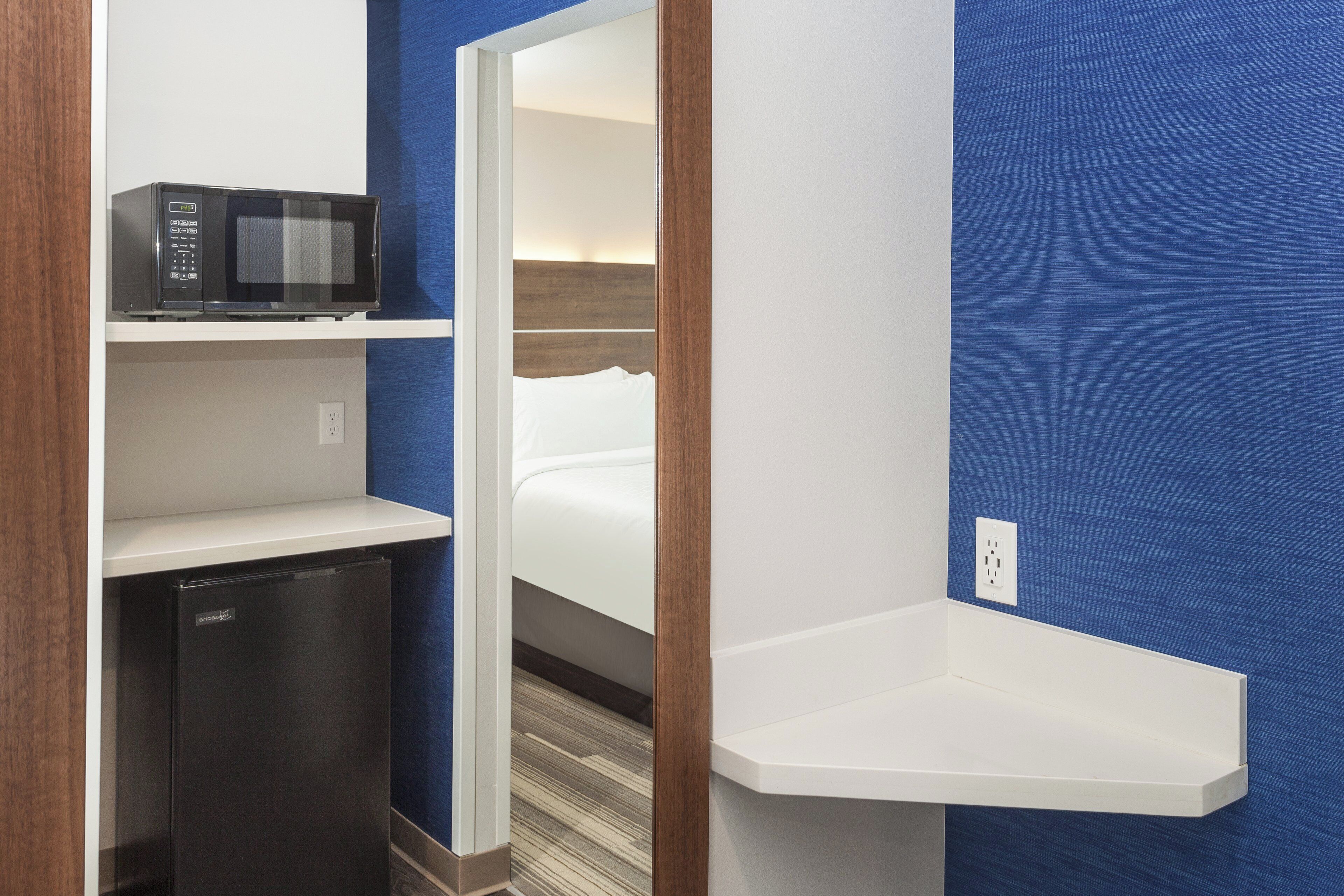 A compact hotel room with a microwave on a shelf, a small refrigerator, and a bedside table.