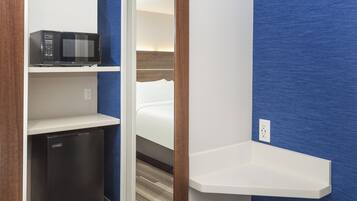 A compact hotel room with a microwave on a shelf, a small refrigerator, and a bedside table.