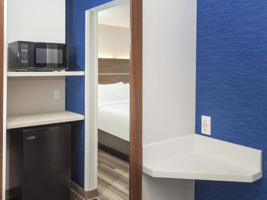 A compact hotel room with a microwave on a shelf, a small refrigerator, and a bedside table.