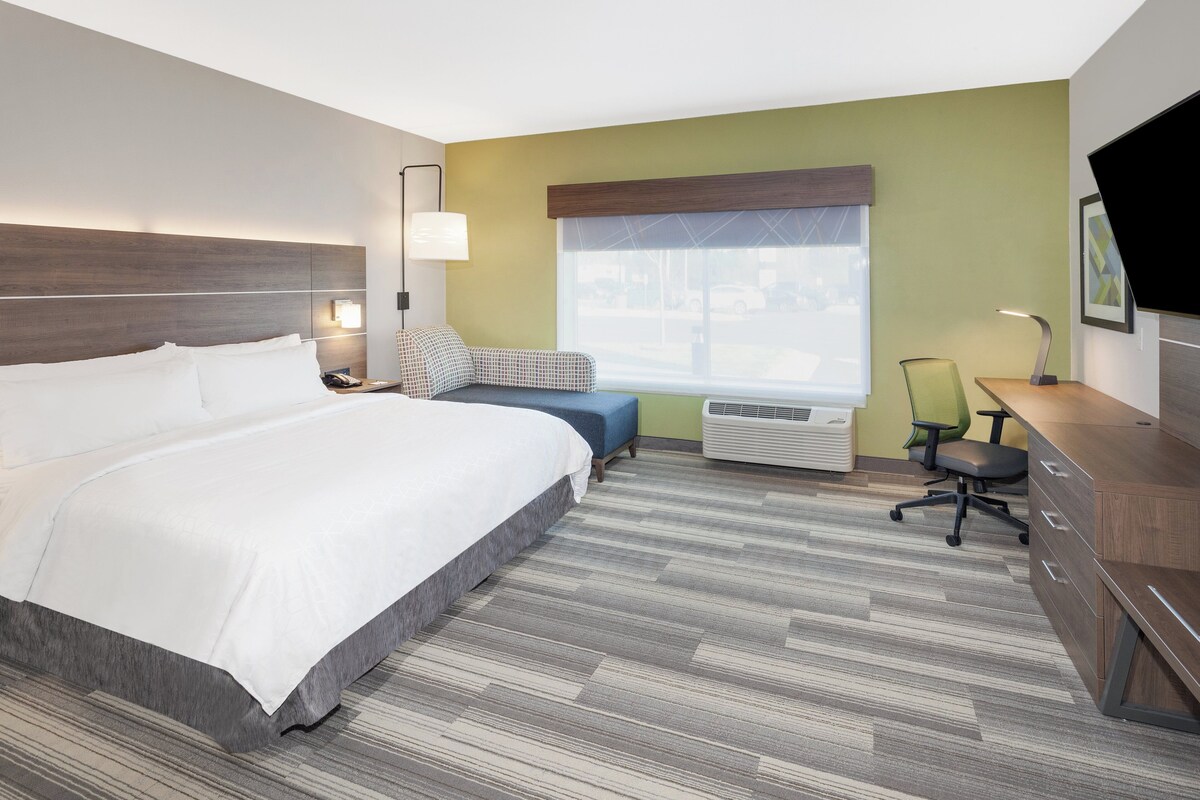 A hotel room with a large bed, a desk, a chair, a window with blinds, an air conditioning unit, and a flat-screen TV.
