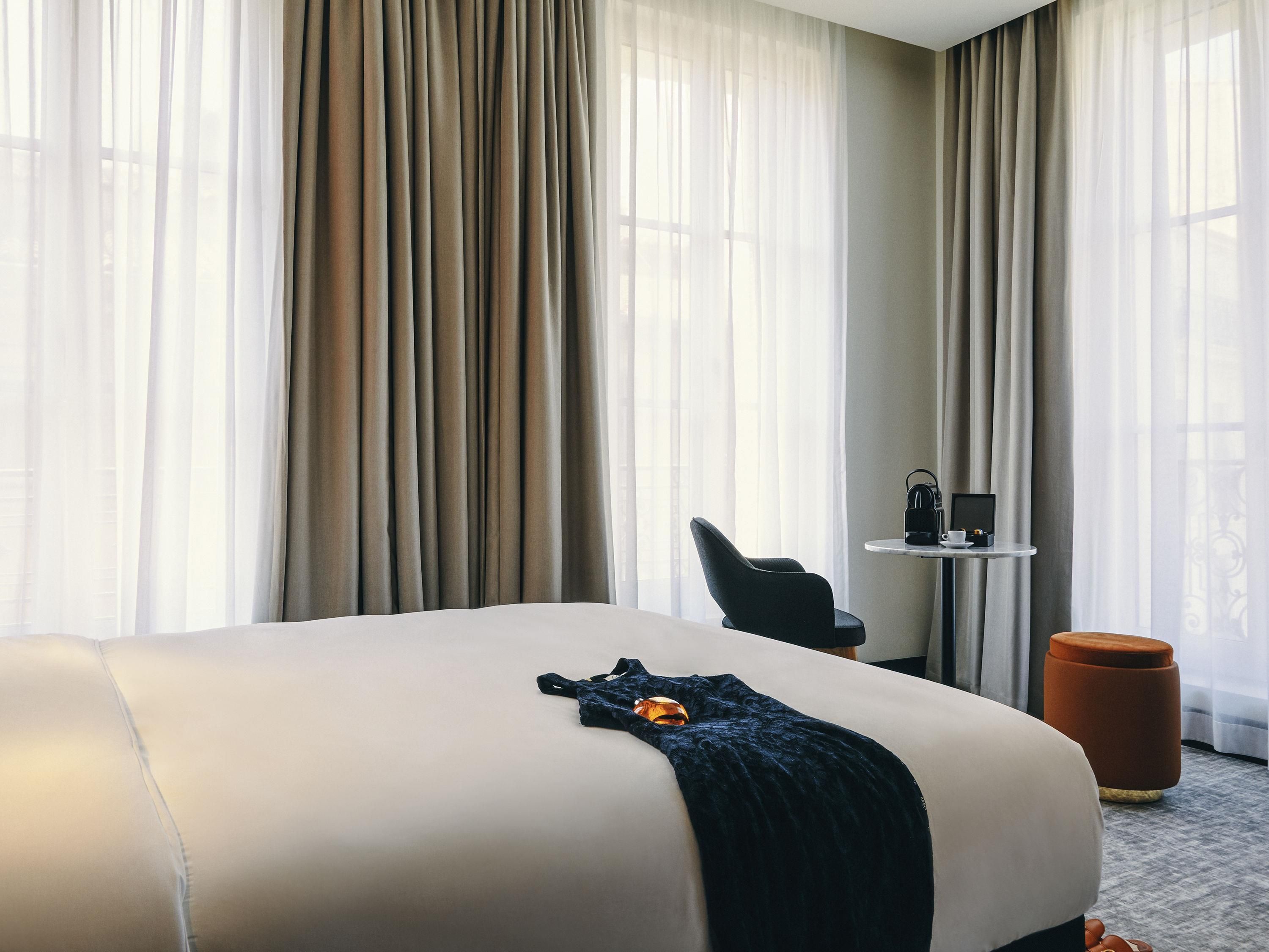 A hotel room with a large bed, a chair, a small table with a coffee maker, and a round ottoman.