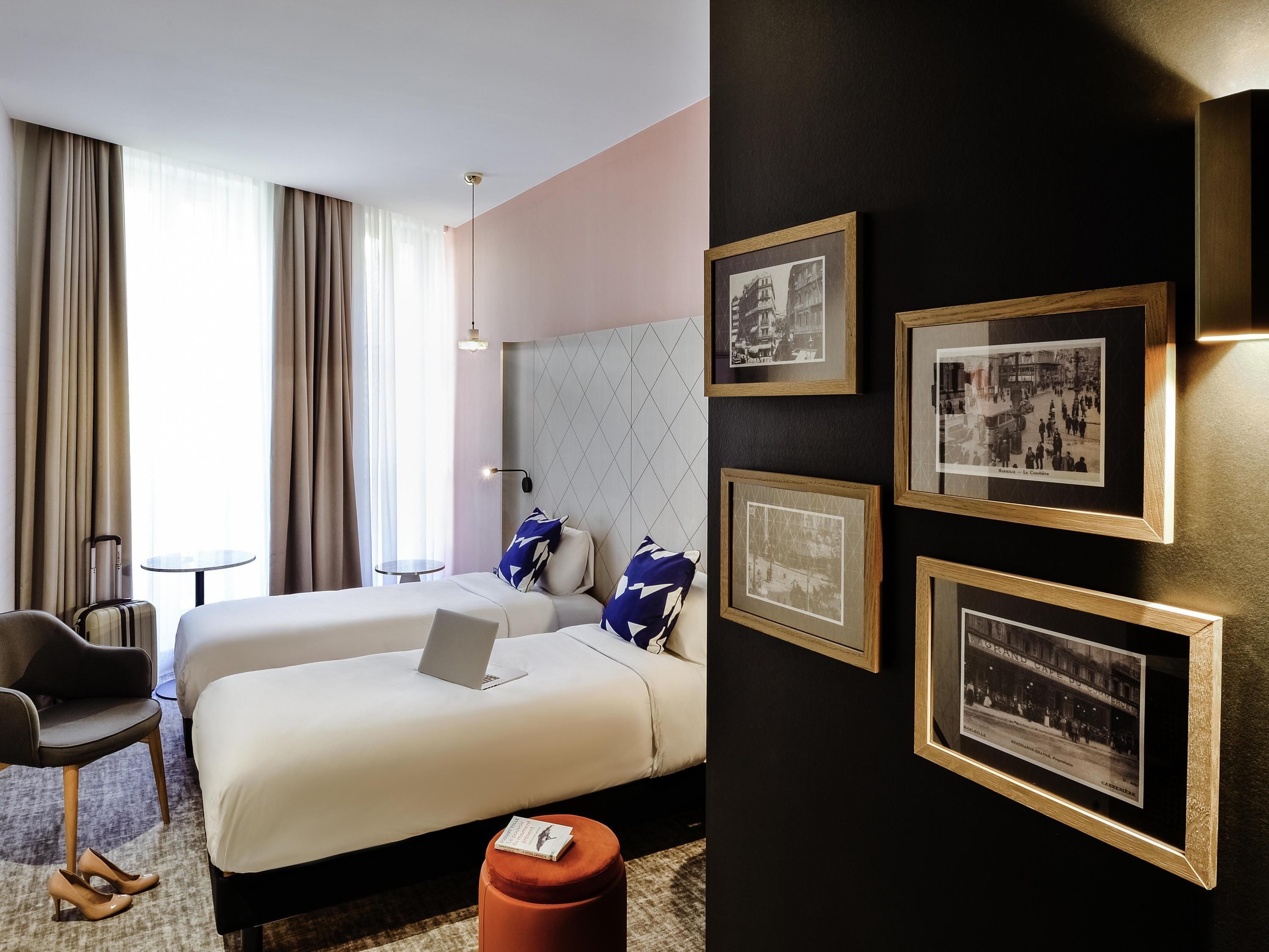 A hotel room with two beds, a desk, a chair, and a small table. There are framed pictures on the wall.