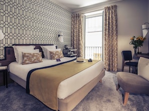 A hotel room with a patterned wall, a bed with multiple pillows, a bedside table with a lamp, a chair, and a view of the outside. - Hôtel Mercure Marseille Canebière Vieux-Port (Marseille)