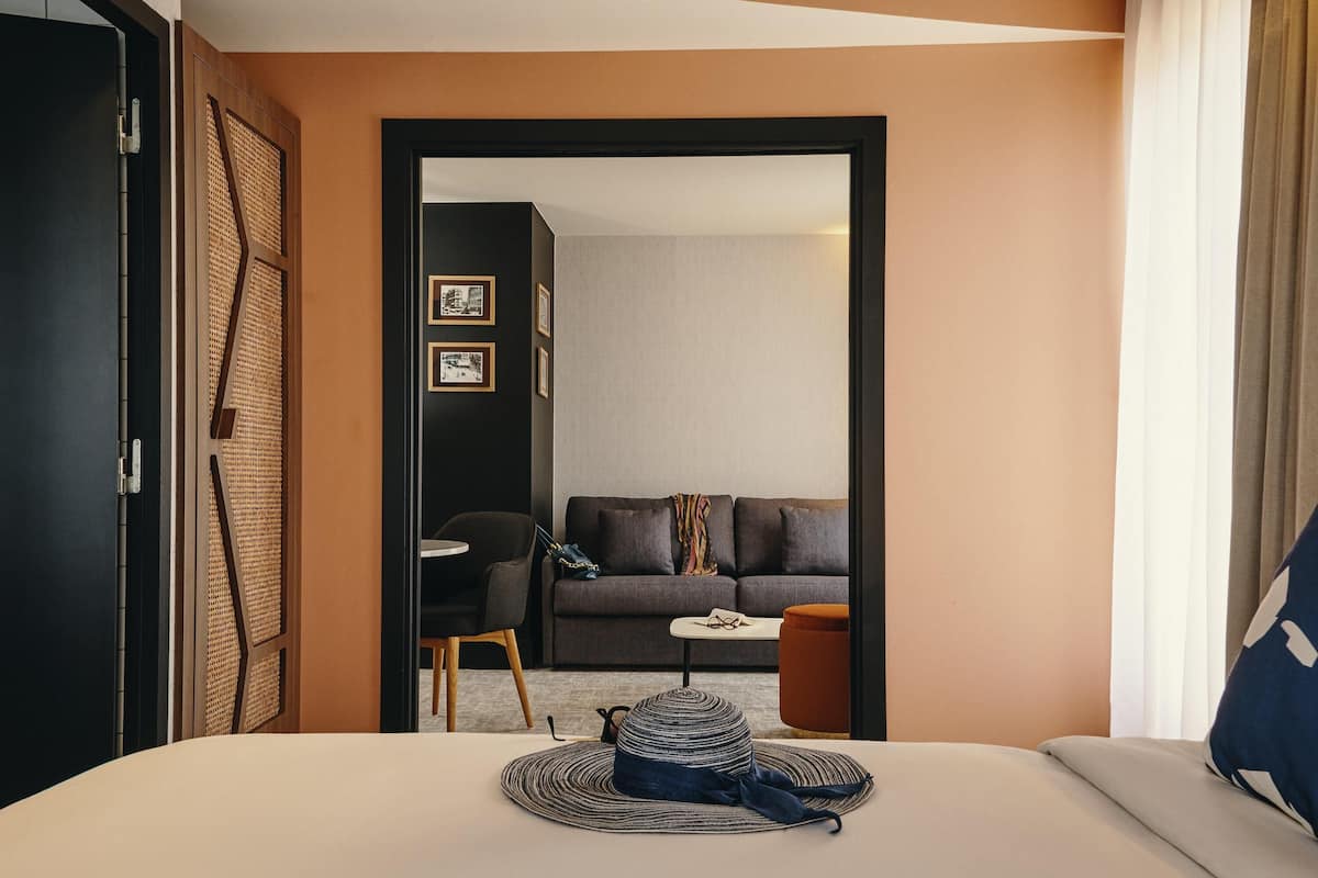 Hotellihuone, jossa on sänky, pieni pöytä, tuoli, sohva ja ovi, joka johtaa toiseen huoneeseen.