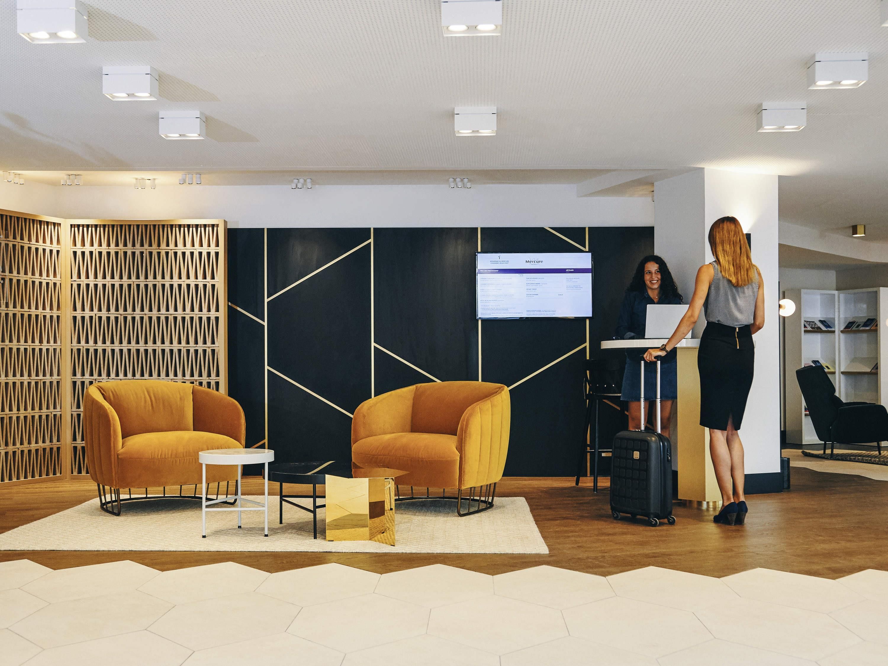 A modern lobby with a reception desk, two yellow armchairs, a white table, and a large screen displaying a menu.