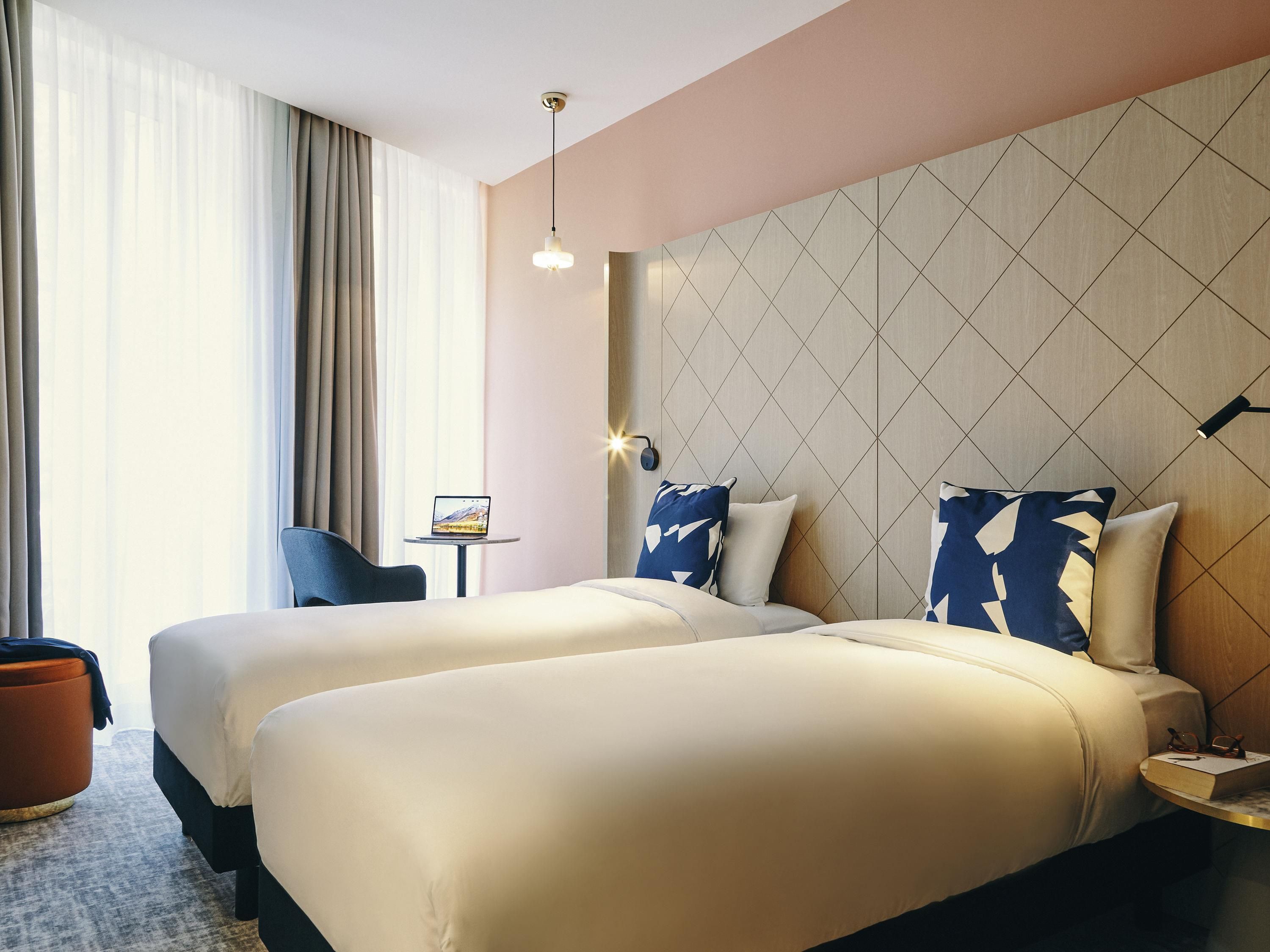 A hotel room with two beds, a small table, a chair, and a large window with curtains.