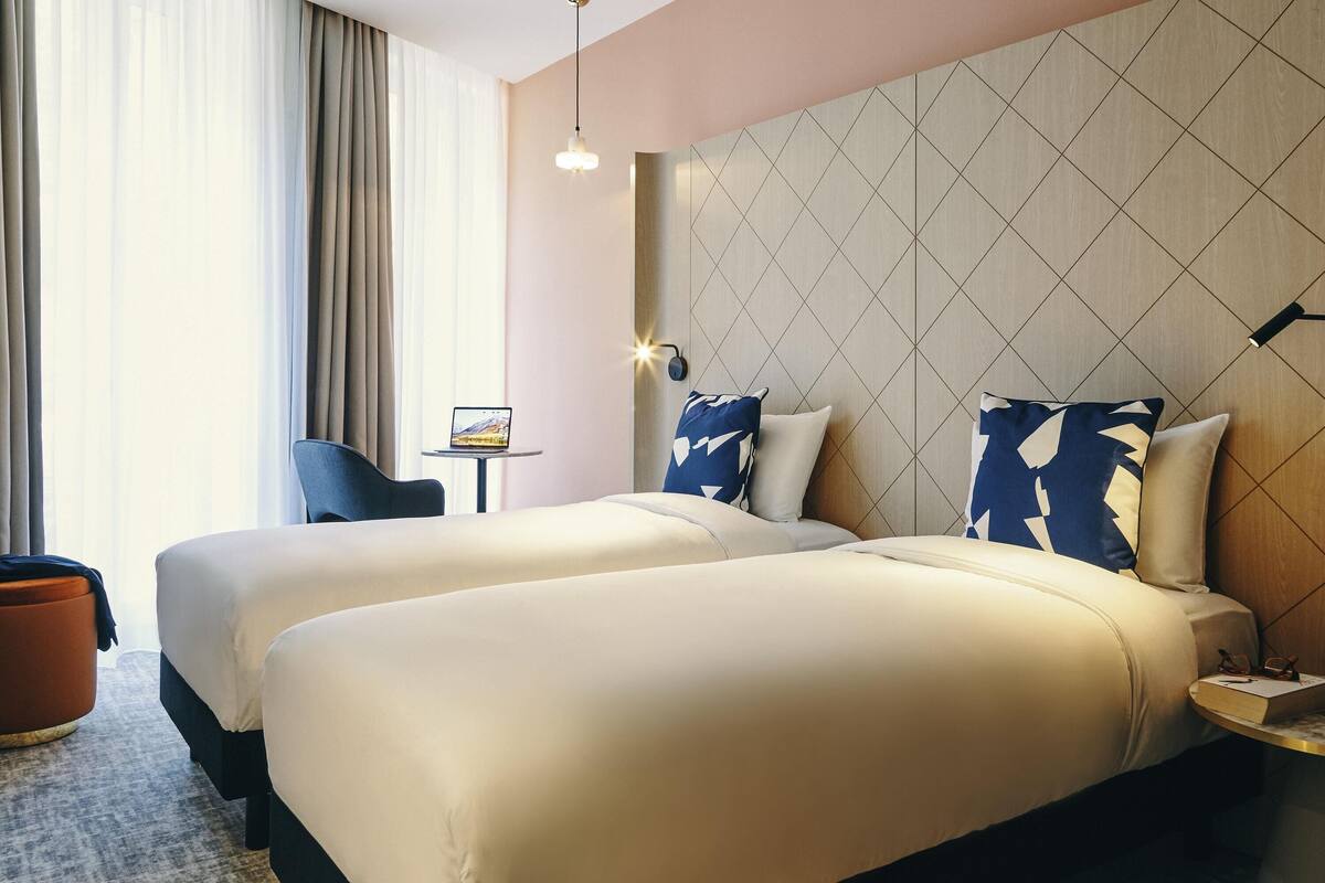 Ein Hotelzimmer mit zwei Betten, einem kleinen Tisch, einem Sessel und einem großen Fenster mit Vorhängen.
