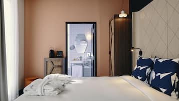 Habitación de hotel con cama, mesita de noche con lámpara, espejo y vistas al baño.