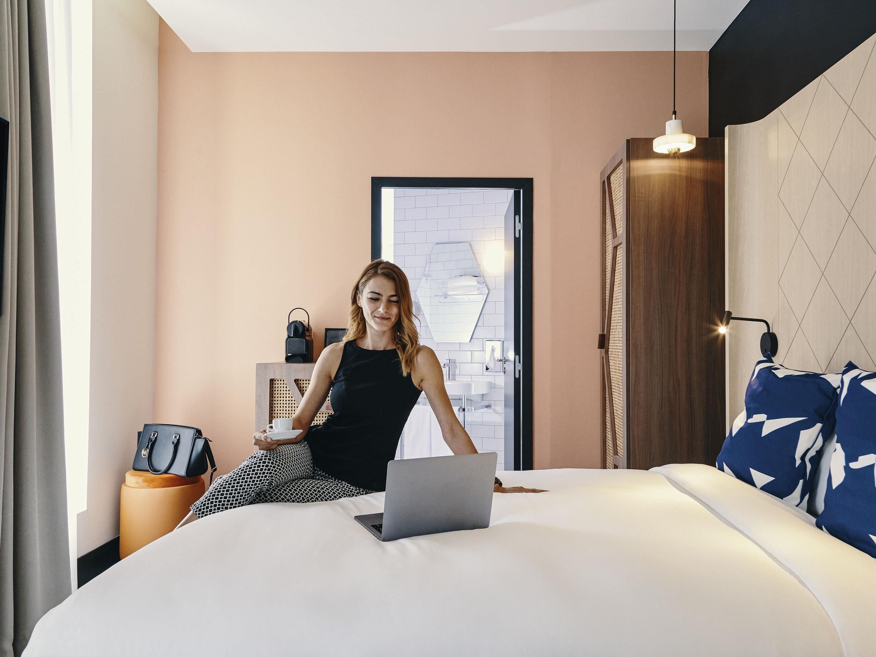Una habitación de hotel moderna con una cama, una computadora portátil, un escritorio con una silla, una maleta y un altavoz instalado en la pared.
