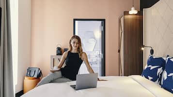Una habitación de hotel moderna con una cama, una computadora portátil, un escritorio con una silla, una maleta y un altavoz instalado en la pared.