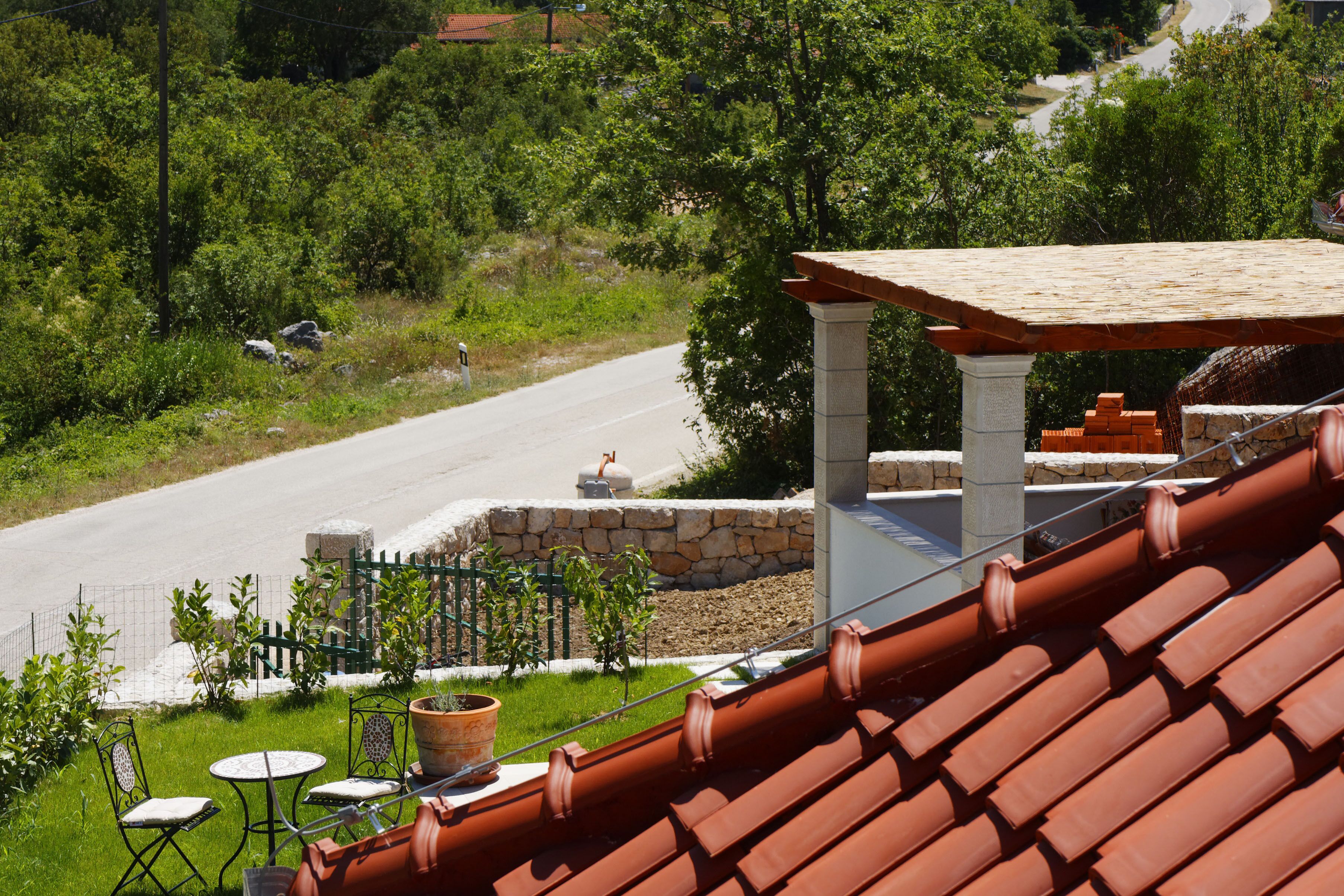 A terrace with a stone wall, a wooden pergola, and a view of a road and greenery.
