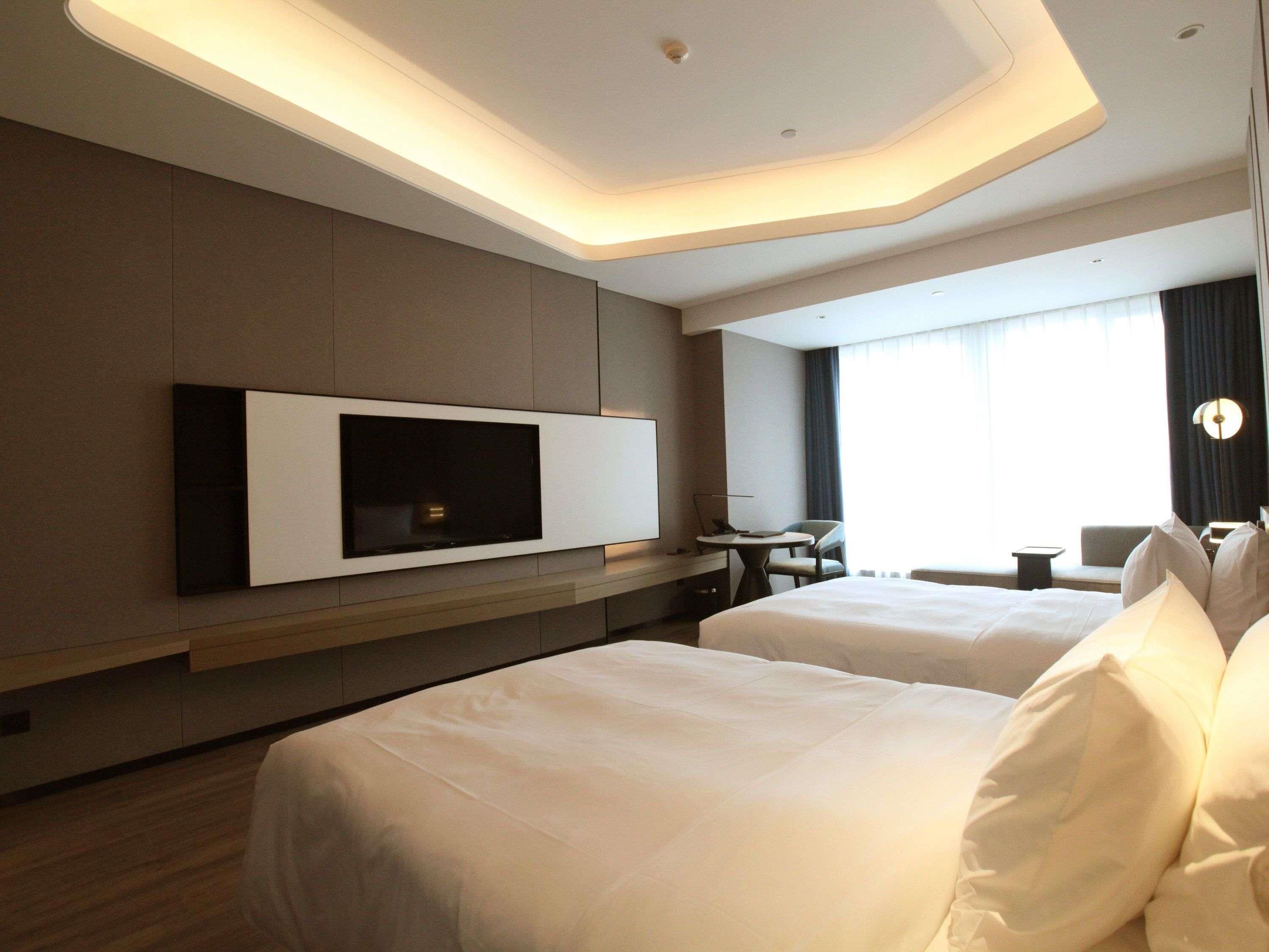 A hotel room with a large flat-screen TV mounted on the wall, two beds with white linens, a small table, and a chair.