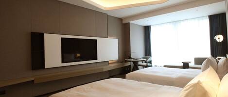 A hotel room with a large flat-screen TV mounted on the wall, two beds with white linens, a small table, and a chair.