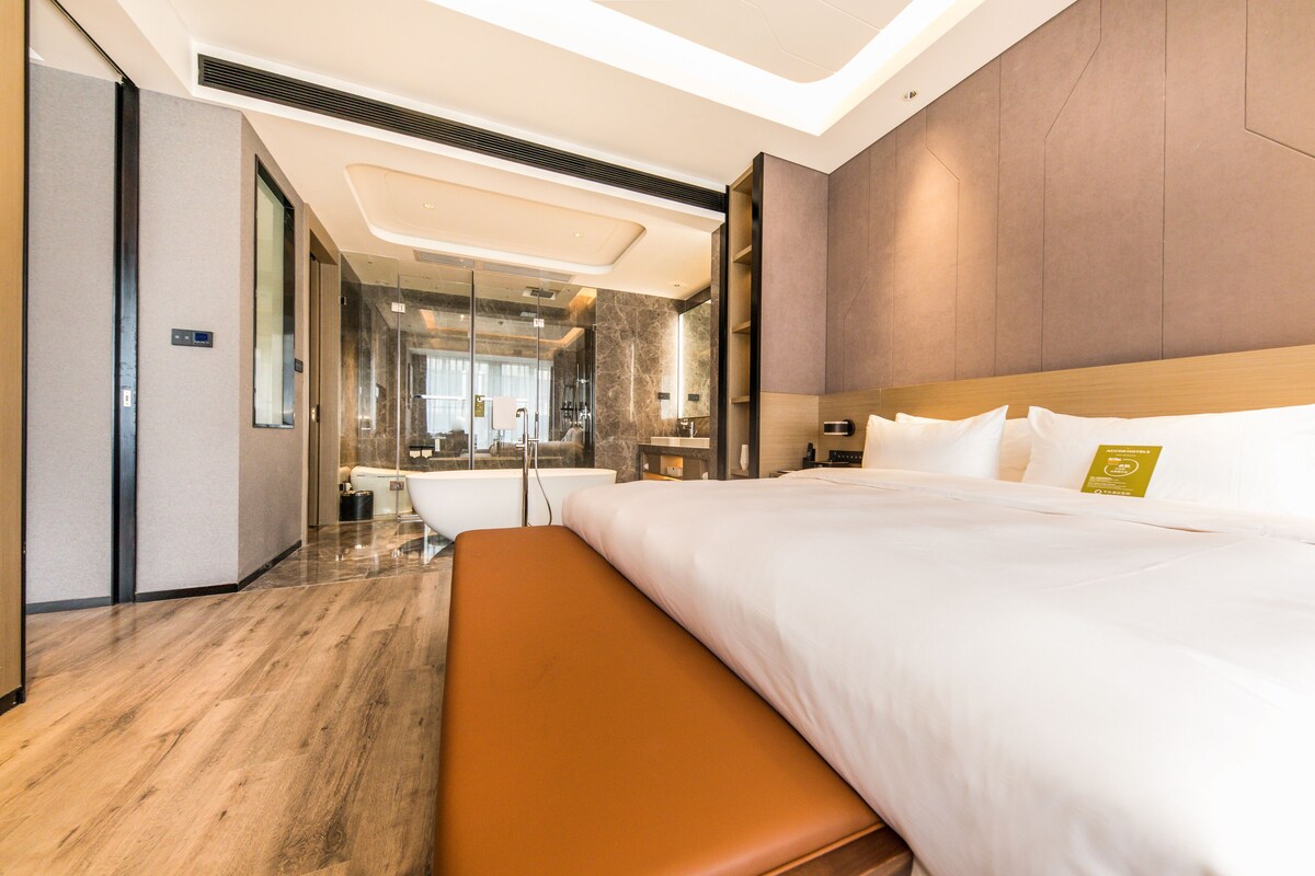 Habitación de hotel moderna con una cama grande, un banco, un baño con paredes de vidrio y vistas a la ciudad.