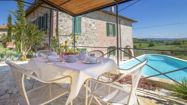 A covered outdoor dining area with a table set for four, overlooking a swimming pool and a stone building with green shutters.