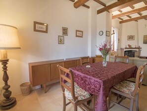 Uma sala de jantar com uma mesa coberta por uma toalha estampada, cadeiras de madeira com almofadas florais, um banco de madeira e um abajur.
