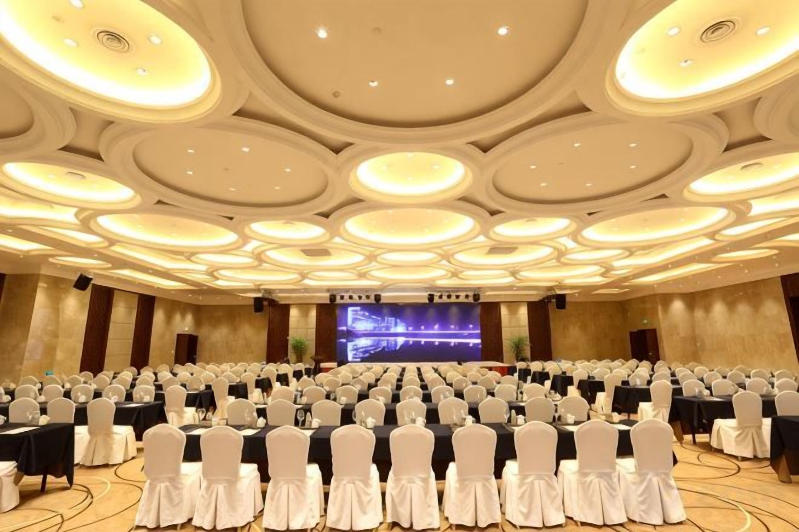 A large conference room with rows of tables and chairs, a stage with a screen, and a modern ceiling design.
