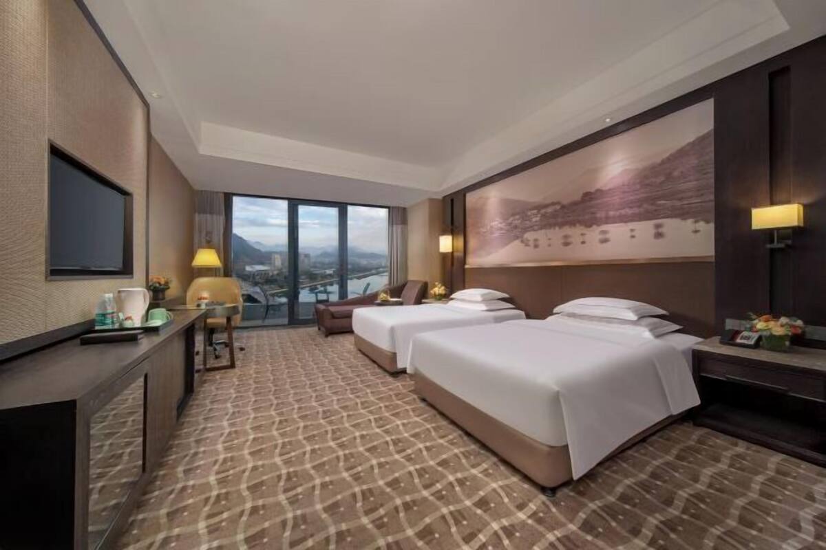 A hotel room with two beds, a desk, a chair, a large window with a city view, and a mounted TV.