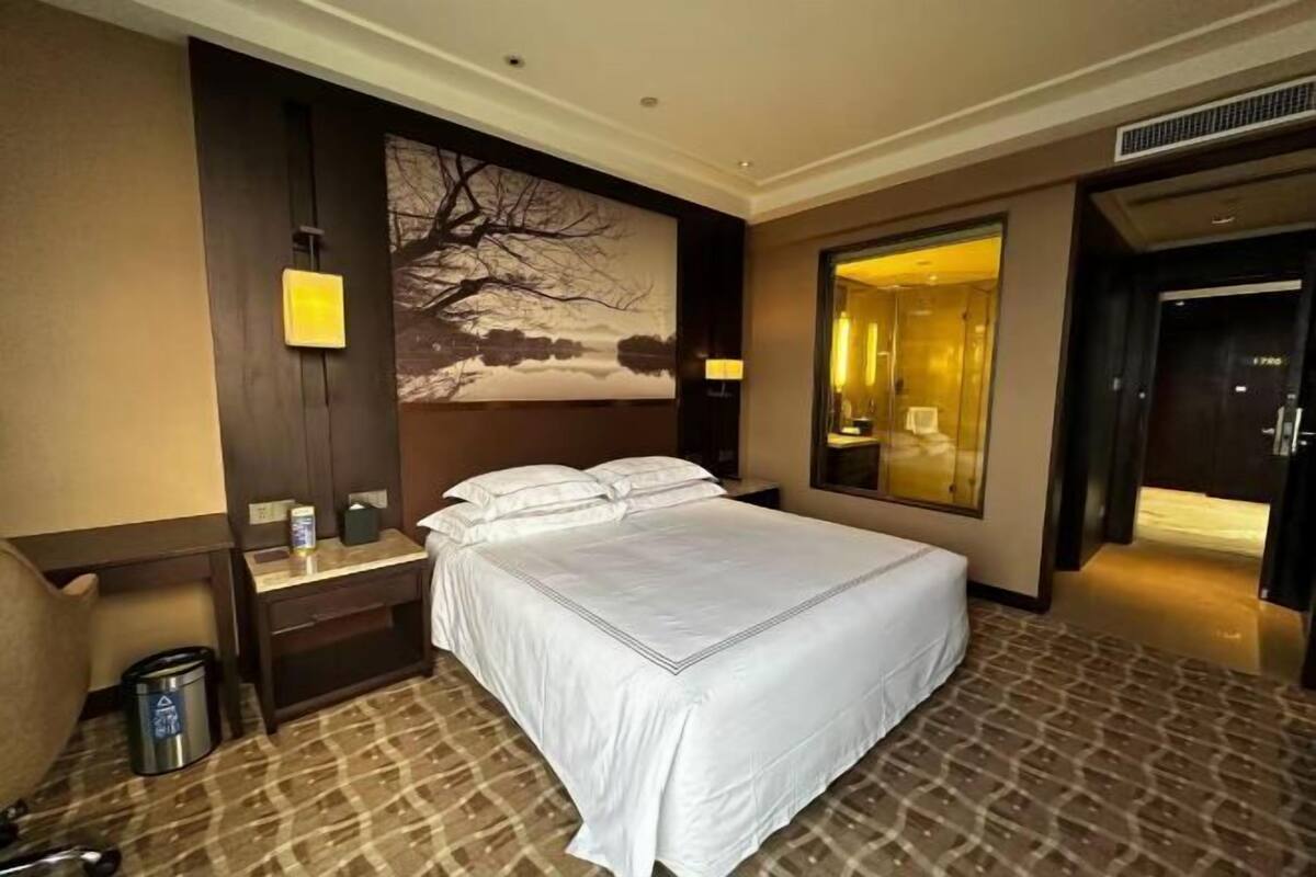 A hotel room with a large bed, a nightstand, a desk, a chair, a trash can, a wall-mounted lamp, and a large window.
