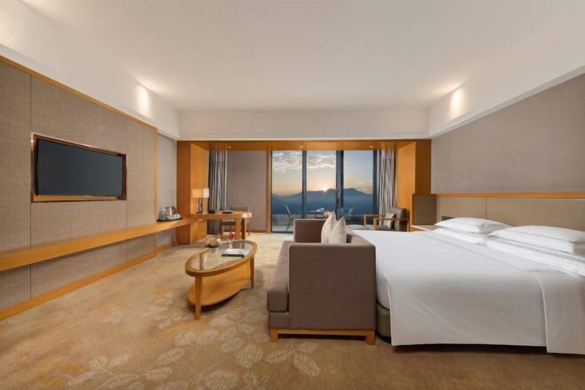 A hotel room with a bed, a sofa, a TV, a small table, and a view of the mountains.