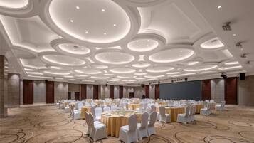 A large conference room with round tables and chairs, a high ceiling with circular light fixtures, and a carpeted floor.