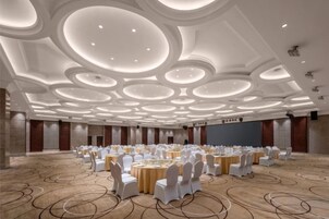Un amplio salón de conferencias con mesas y sillas redondas, un techo alto con luminarias circulares y suelo alfombrado.