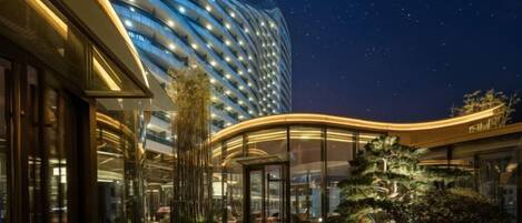 A hotel with a modern curved facade, a well-lit entrance, and a landscaped garden area.