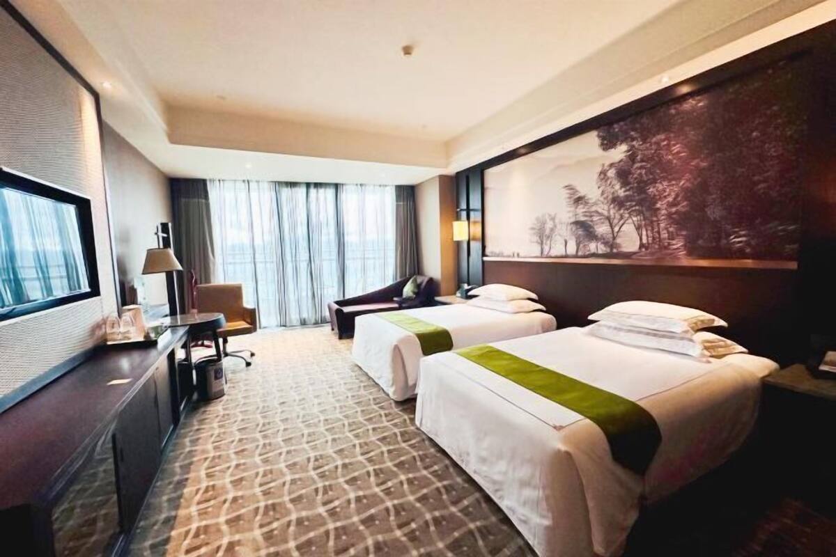A hotel room with two beds, a desk, a chair, a TV, and a large painting on the wall.