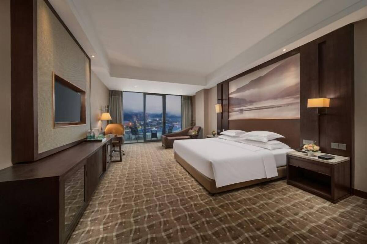 A hotel room with a large bed, a desk, a chair, a view of the city through large windows, and a painting on the wall.