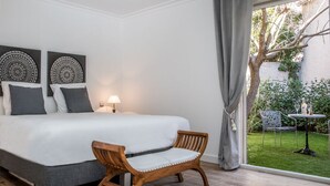 Ein Schlafzimmer mit einem Bett, zwei Wandbildern, einem Holzstuhl, einem Nachttisch und einem Fenster mit Blick auf einen Garten und Gartenmöbel.
