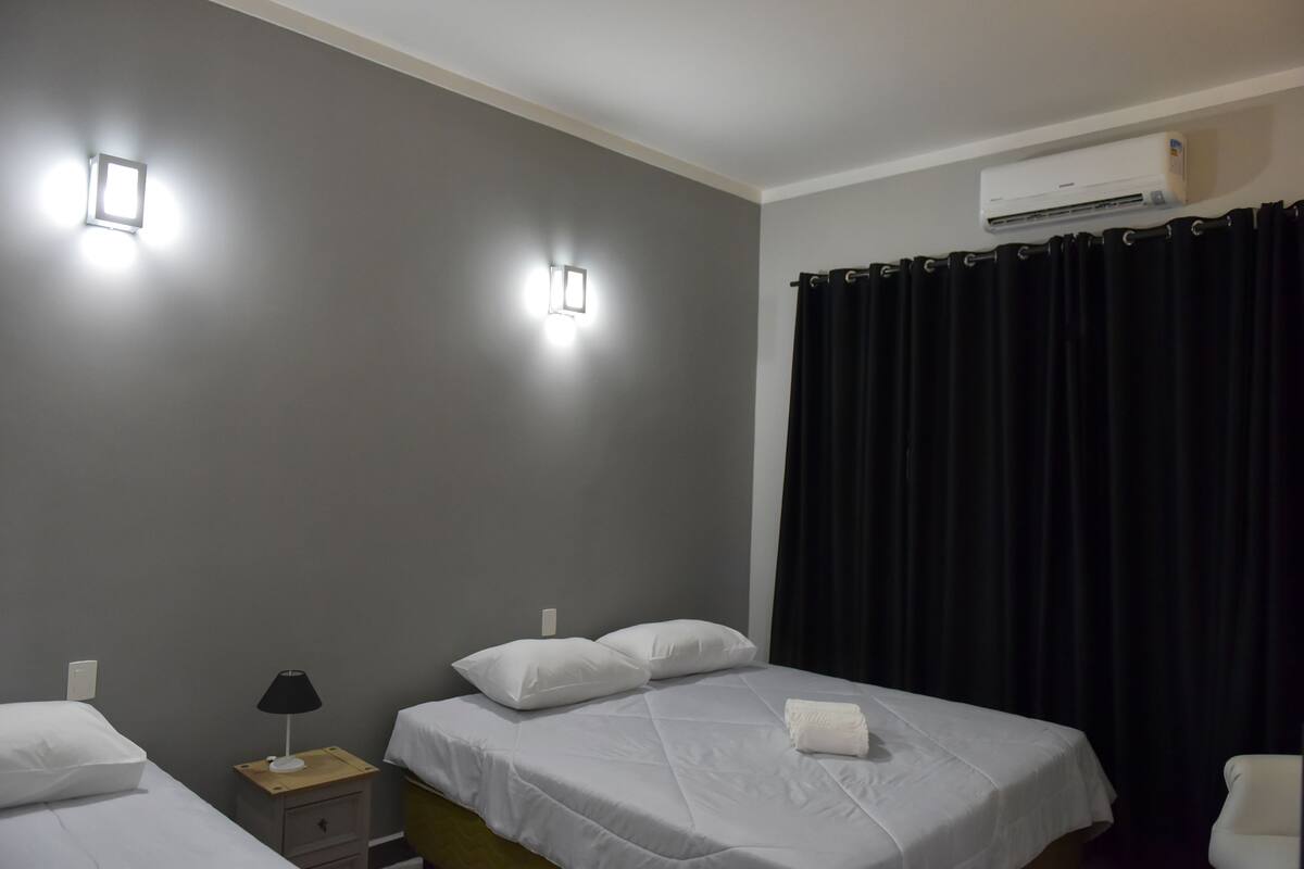 Habitación de hotel con dos camas, una mesita de noche con lámpara, aire acondicionado instalado en la pared y cortina negra.