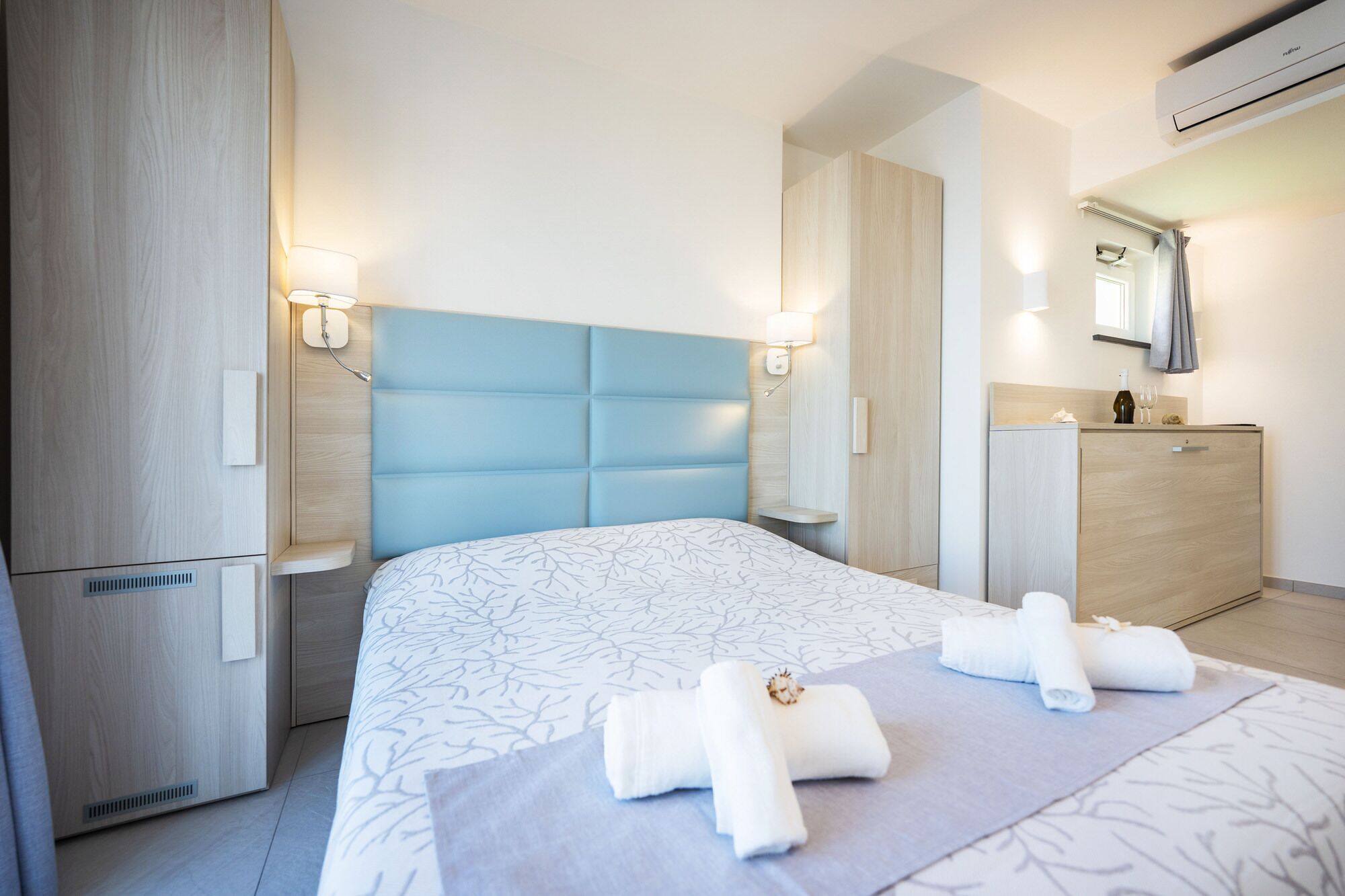 Una habitación de hotel moderna con una cama grande, un armario de madera, un tocador con espejo y un baño a la vista al fondo.